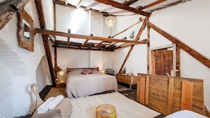Een rustieke slaapkamer met zichtbare houten balken, een groot bed, een houten dressoir en een geweven poef.