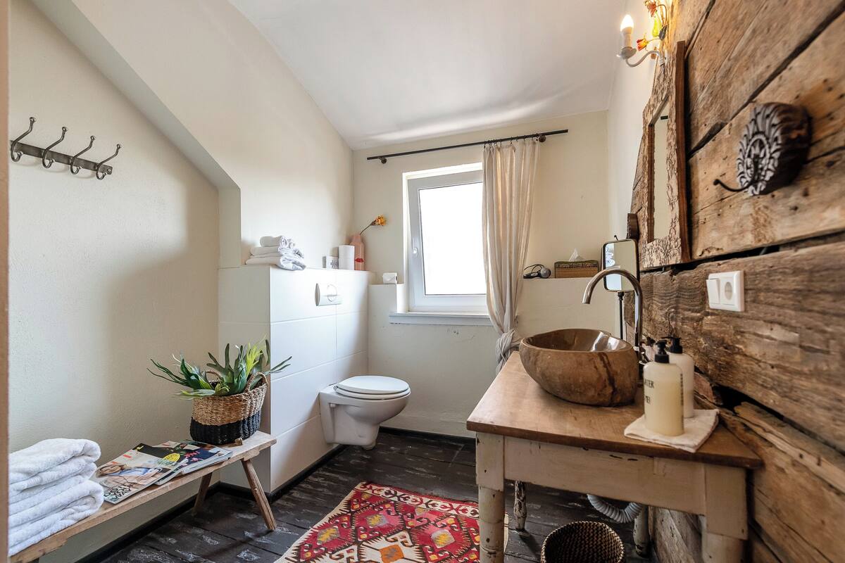 A bathroom with a wooden vanity, a sink, a toilet, a window with curtains, a shelf with toiletries, and a plant on a shelf.