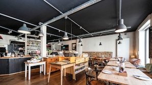 Een ruime restaurantruimte met hoge plafonds, zichtbare balken en hanglampen. Tafels staan klaar voor diners, en de keuken is op de achtergrond zichtbaar.