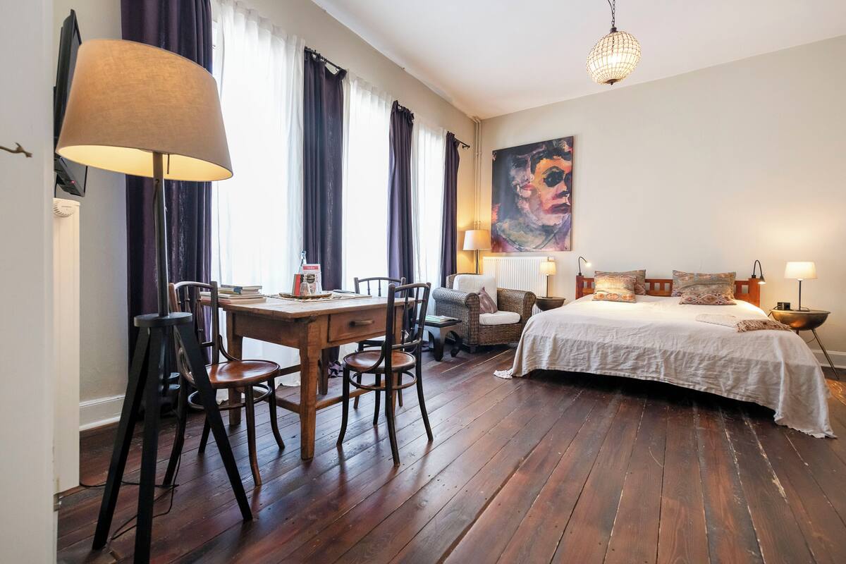 A room with a wooden dining table, chairs, a bed with a white bedspread, a large painting on the wall, and a hanging light fixture.