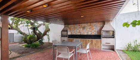 An outdoor patio with a brick floor, a wooden pergola, a table with chairs, and a built-in grill area.