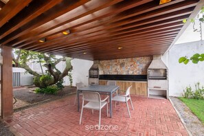 An outdoor patio with a brick floor, a wooden pergola, a table with chairs, and a built-in grill area. - House in Condo w/ Pool and BBQ Nto0005 (Camaçari)