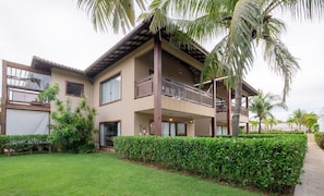 A two-story building with a balcony, surrounded by greenery and palm trees. - House in Condo w/ Pool and BBQ Nto0005 (Camaçari)