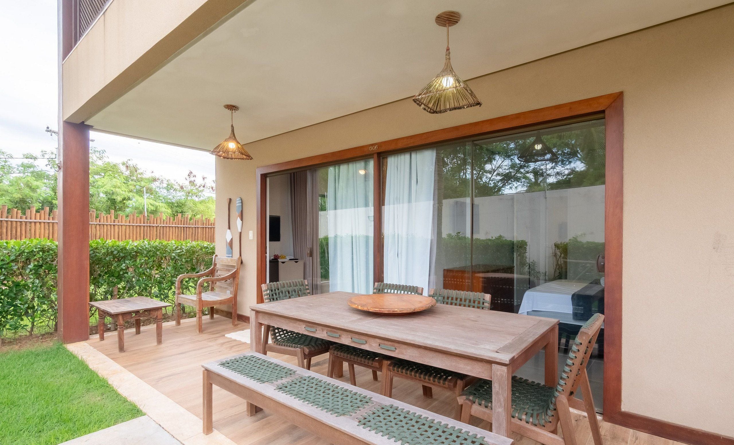 Un patio exterior con mesa y bancos de madera, una valla de madera y un ventanal de vidrio que da a un área techada.