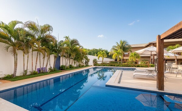 A swimming pool with clear blue water, surrounded by palm trees and a covered patio area with umbrellas. - House in Condo w/ Pool and BBQ Nto0005 (Camaçari)