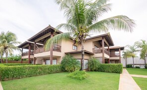 A two-story building with balconies, surrounded by well-maintained lawns and palm trees. - House in Condo w/ Pool and BBQ Nto0005 (Camaçari)