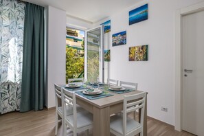 Un coin repas avec une table dressée pour quatre personnes, des chaises blanches, une fenêtre donnant sur des bâtiments et de la verdure, et une porte sur la droite.