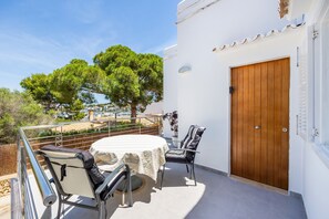 En balkon med et bord og stole, en trädør og udsigt til træer og en strand.