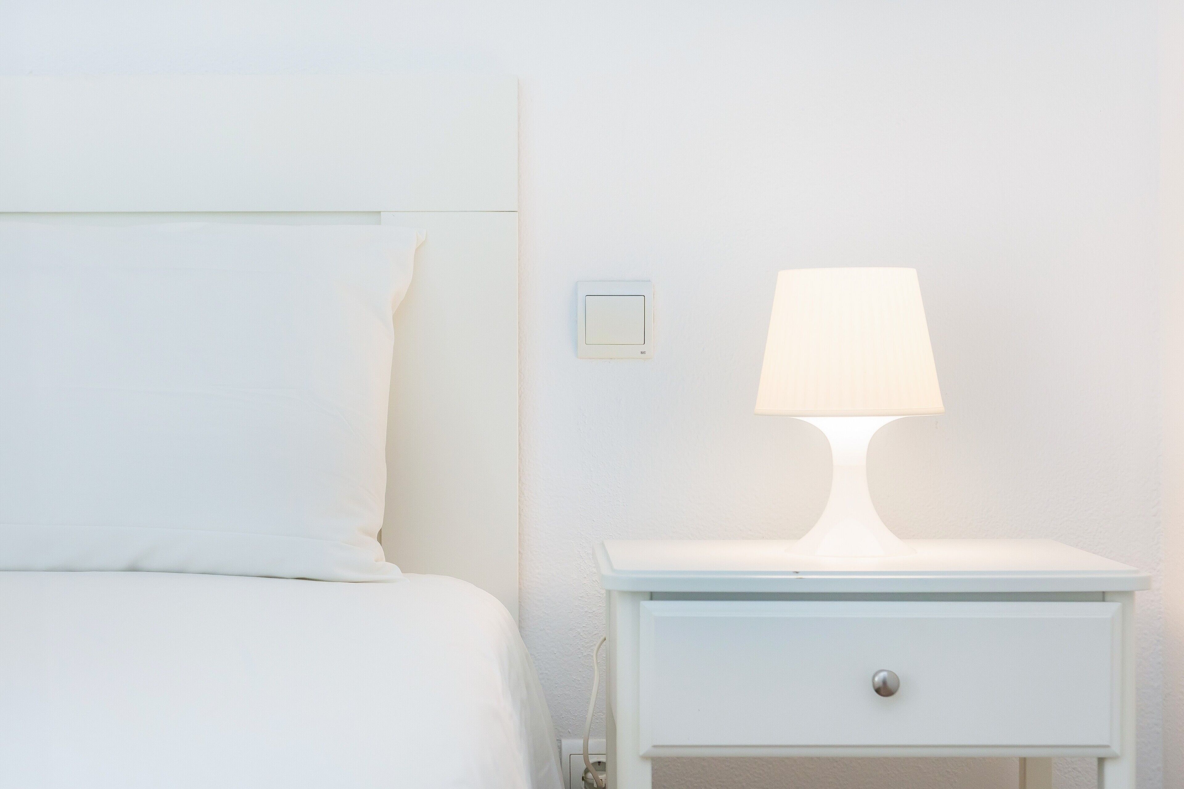 A white bed with a pillow, a white nightstand with a lamp, and a wall switch.