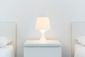 Uma mesa de cabeceira branca com uma luminária, uma parede branca e uma cama com roupa de cama branca.