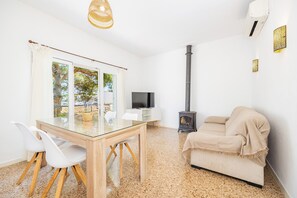 Uma sala de estar moderna com uma mesa de jantar de vidro, cadeiras brancas, um sofá bege, uma TV, uma lareira e uma janela grande com cortinas.