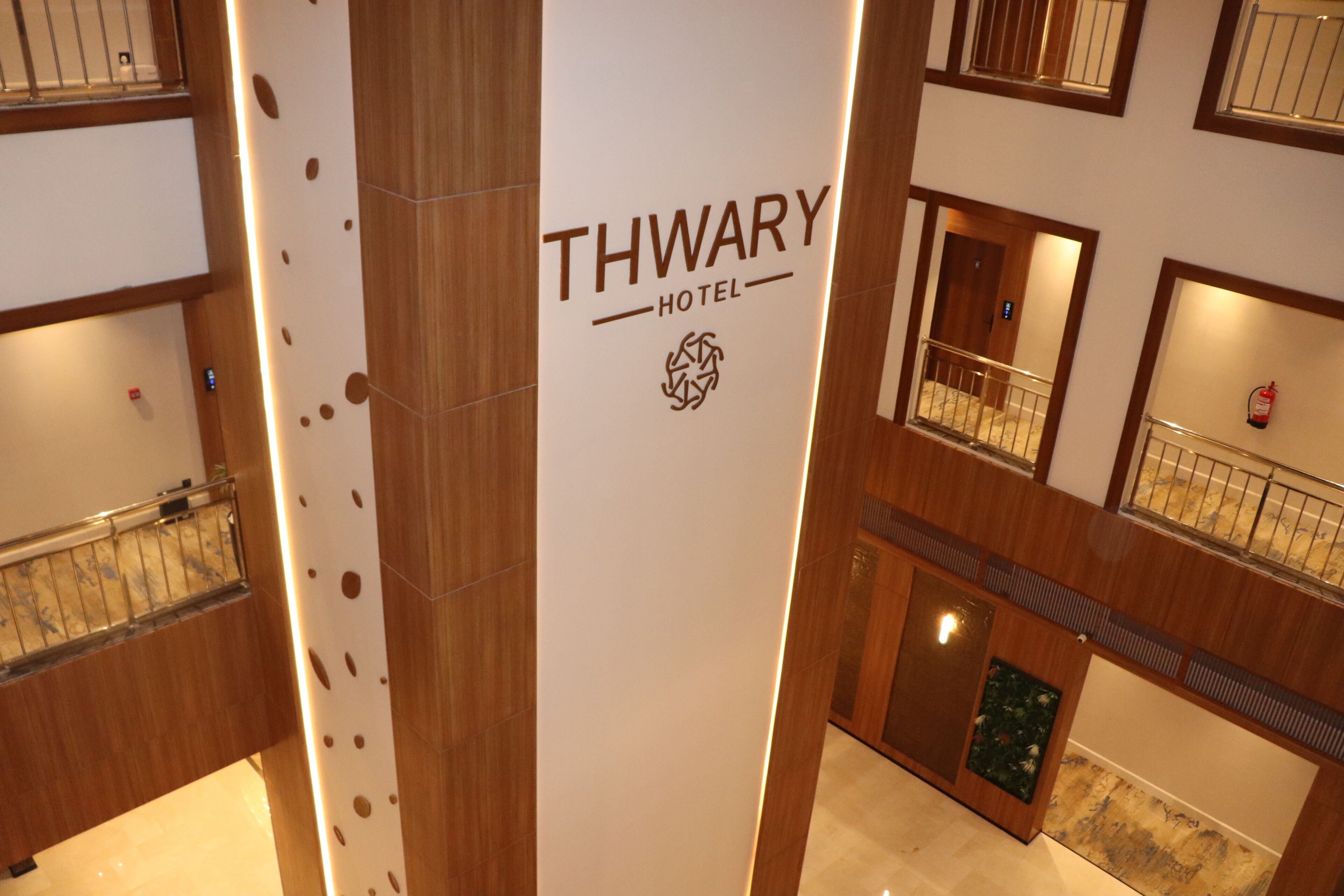 Een THWARY HOTEL bord met een logo, gelegen in een gebouw met meerdere verdiepingen, houten lambrisering en een glazen balustrade.
