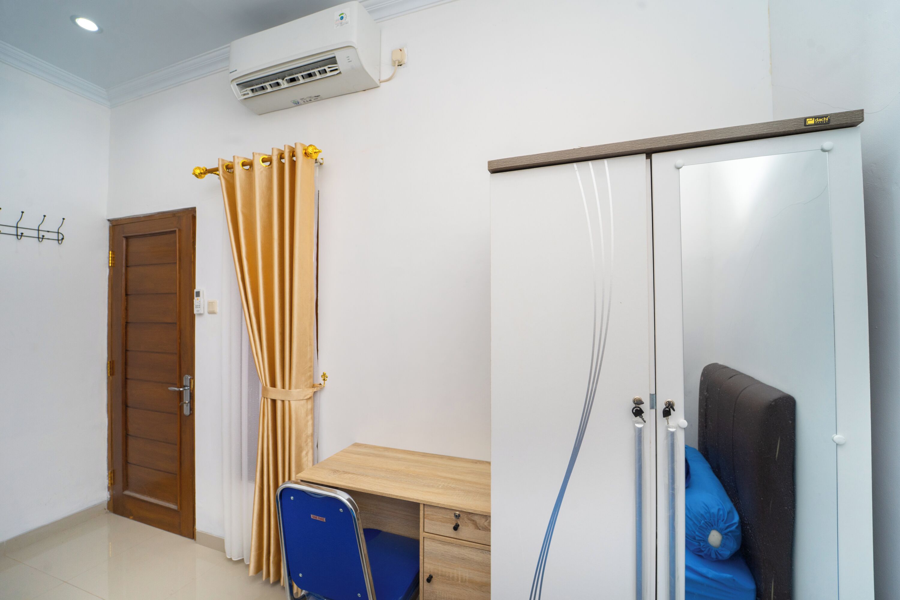 A room with a wooden door, beige curtains, a blue chair, a wooden desk, and a wardrobe with a mirror and a black headboard.