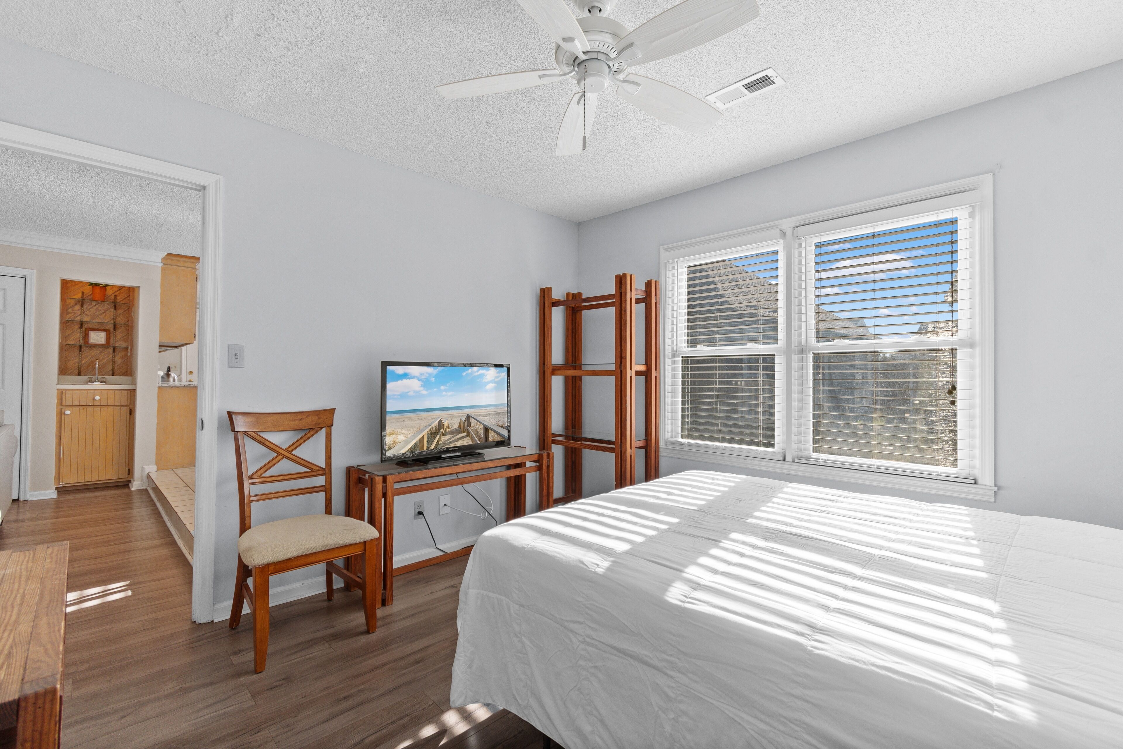 A bedroom with a bed, a TV on a wooden stand, a chair, and a window with blinds.