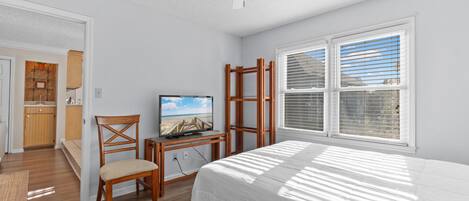 A bedroom with a bed, a TV on a wooden stand, a chair, and a window with blinds.