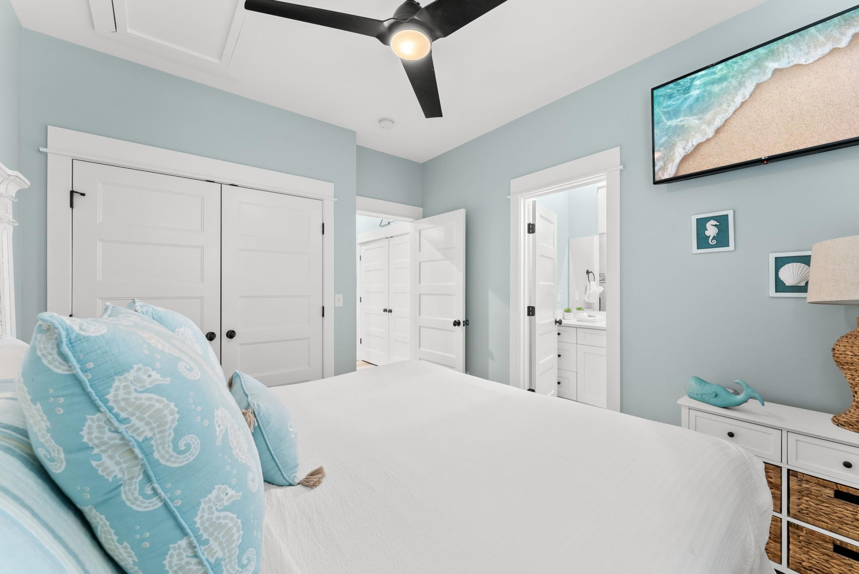 Une chambre à coucher avec un lit, une armoire blanche, un ventilateur de plafond et un téléviseur fixé au mur qui diffuse une scène de plage.