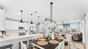 A modern kitchen with white cabinets, a central island, pendant lighting, and a dining area with a wooden table and chairs. - Pet Friendly Beach House - Seminole Wind (Port St. Joe)
