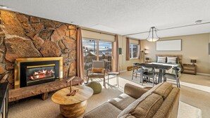A living room with a stone wall, a fireplace, a dining area, and a large window. - Cute, cozy studio at Storm Meadows~Ski In, Walk Out! (Steamboat Springs)