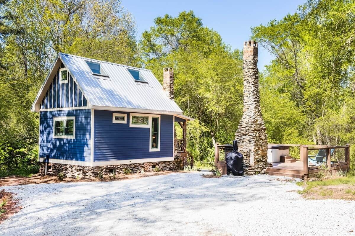 Una pequeña casa de un piso con techo metálico inclinado, chimenea de piedra y camino de entrada de grava.