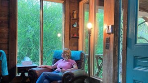 A cozy cabin interior with a wooden floor, a blue door, and a person relaxing on a leather chair by a window.
