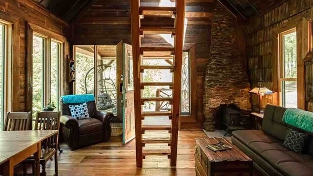 A rustic cabin interior with wooden floors, a stone fireplace, a ladder leading to a loft, and a large rug.