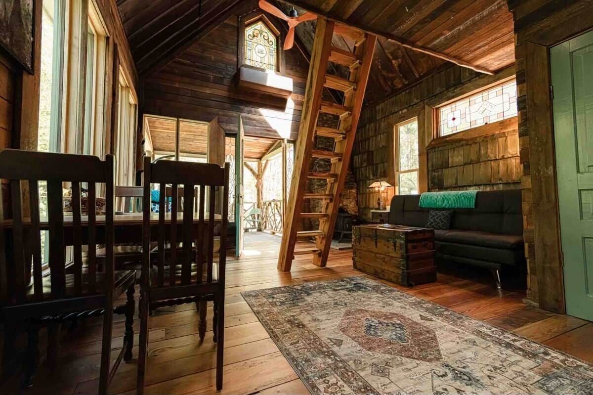Interior rústico de uma cabana com piso de madeira, escada, sofá e mesa de jantar.