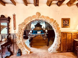 A bedroom with a stone archway, a bed with blue bedding, a wooden dresser, and a nightstand with a lamp.