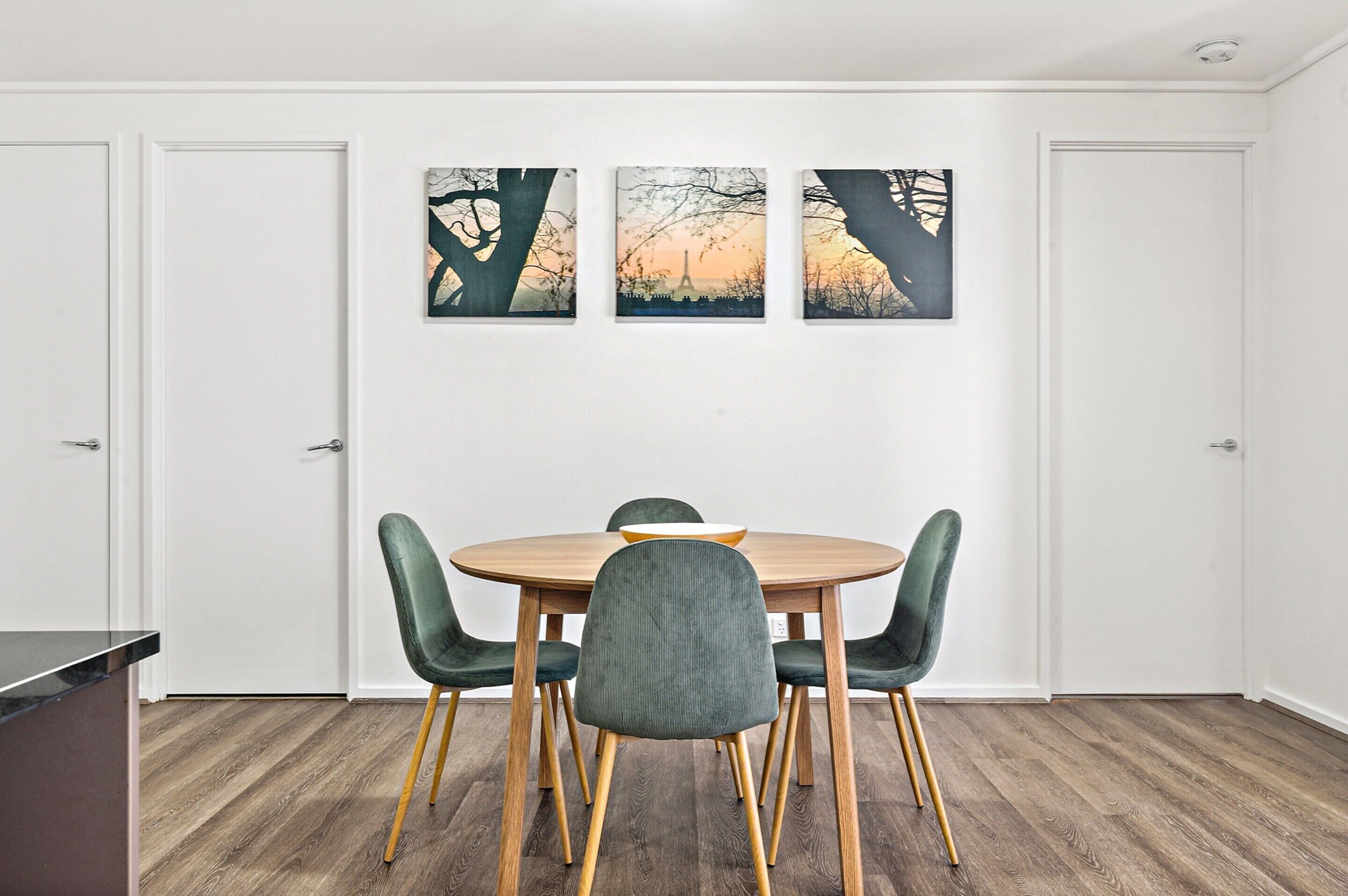 A dining area with a round wooden table, four green upholstered chairs, and three framed artworks depicting trees and a sunset.
