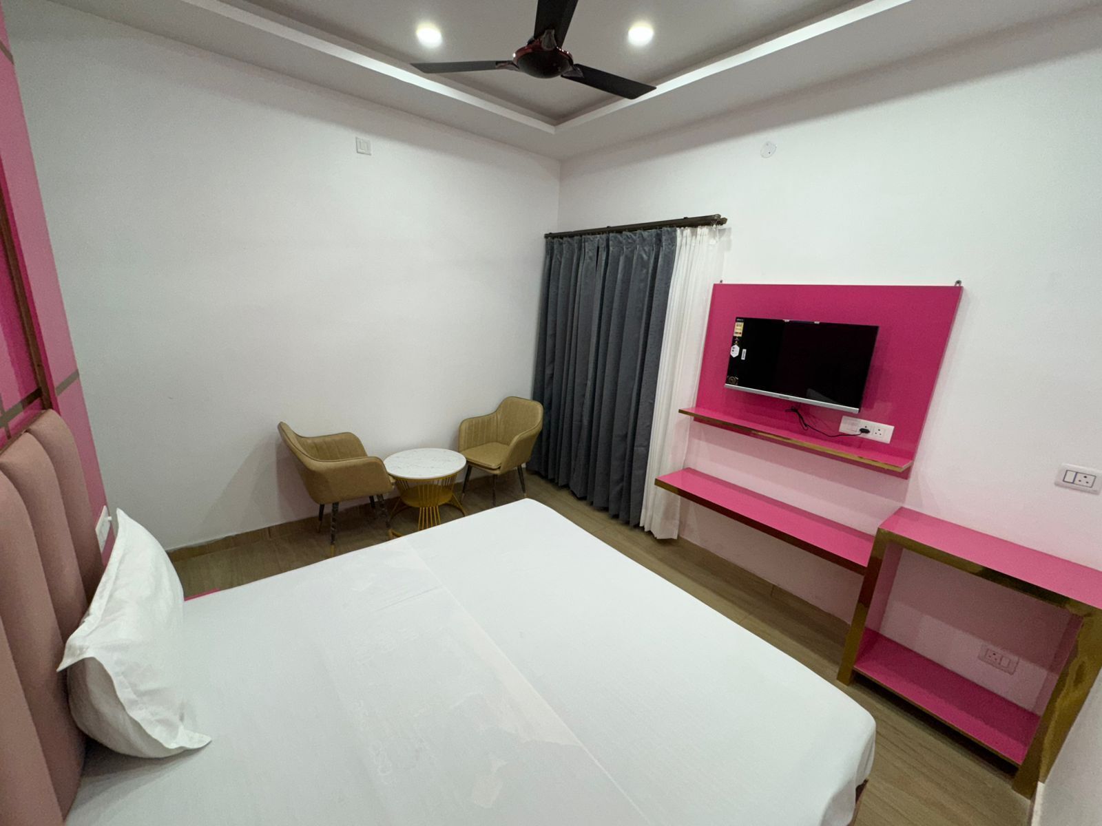 A hotel room with a bed, a TV mounted on a pink wall, a ceiling fan, and two chairs around a small table.