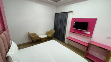 A hotel room with a bed, a TV mounted on a pink wall, a ceiling fan, and two chairs around a small table.