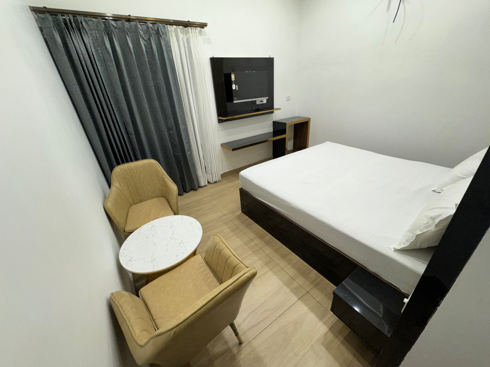 Een kleine hotelkamer met een bed, een klein rond tafeltje, een stoel, een aan de muur bevestigde televisie en een gordijn.