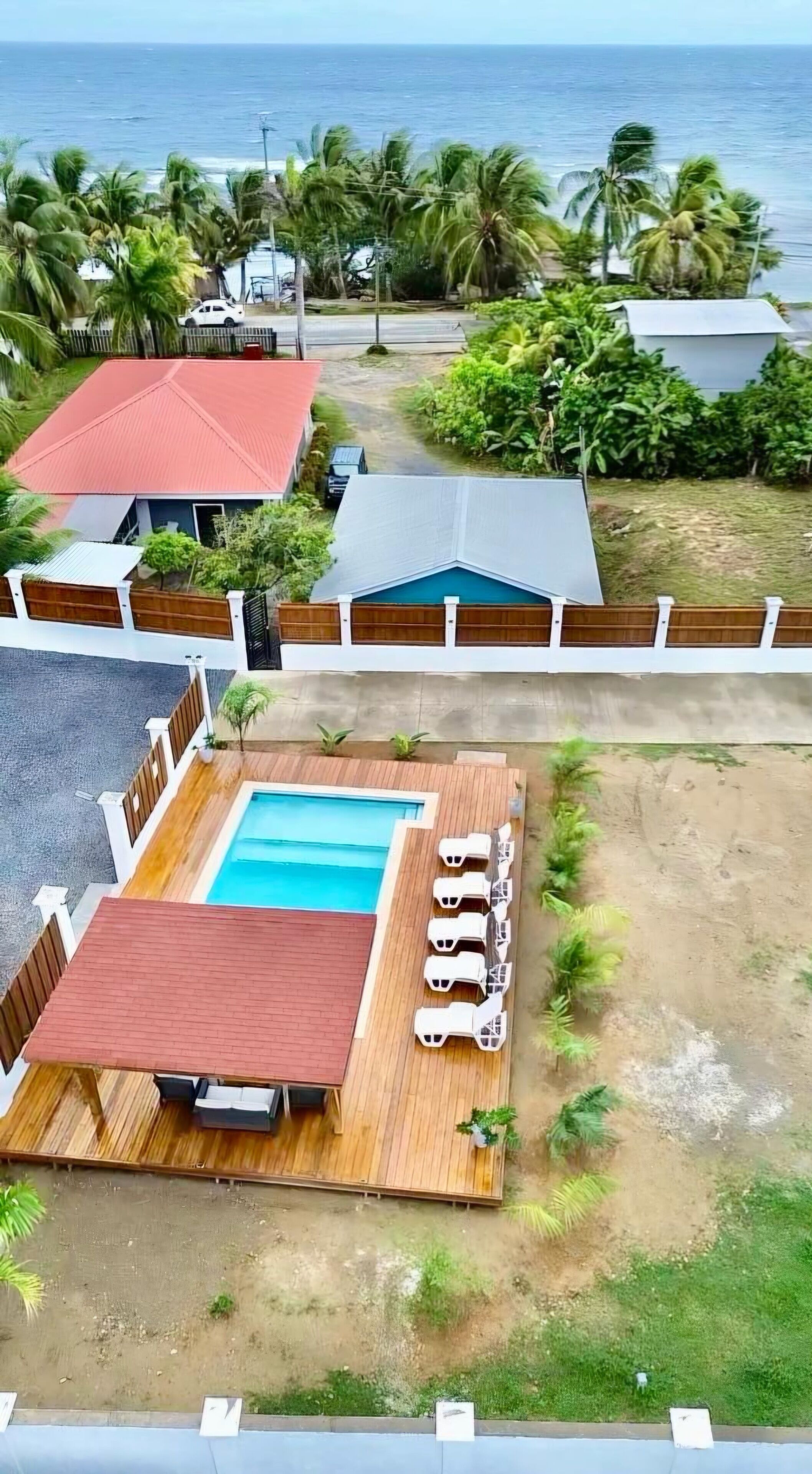 A pool area with a wooden deck and lounge chairs, surrounded by a fence and palm trees, with a view of the ocean.