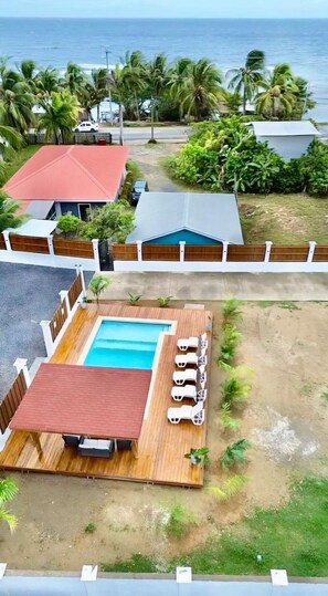 A pool area with a wooden deck and lounge chairs, surrounded by a fence and palm trees, with a view of the ocean.