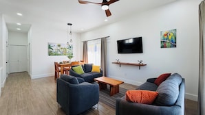 A modern living room with a ceiling fan, a flat-screen TV, a dining area with wooden furniture, and a painting on the wall.