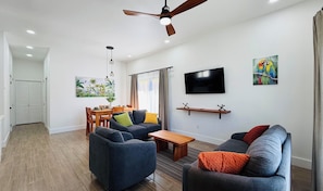A modern living room with a ceiling fan, a flat-screen TV, a dining area with wooden furniture, and a painting on the wall.