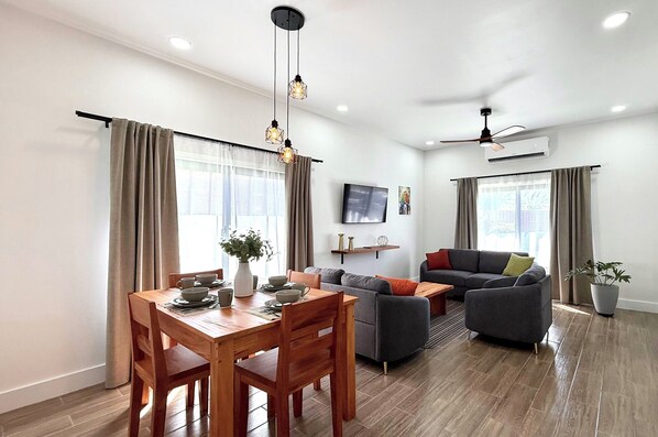 A modern living room with a sofa, dining table, and chairs. There is a ceiling fan and an air conditioner.