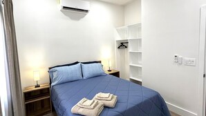 A modern bedroom with a bed, bedside tables, a ceiling fan, an air conditioning unit, and a wardrobe.