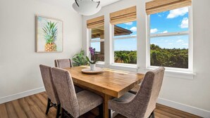 Comedor con mesa de madera y cuatro sillas, ventanas amplias con vistas a vegetación y una obra de arte con forma de piña en la pared.