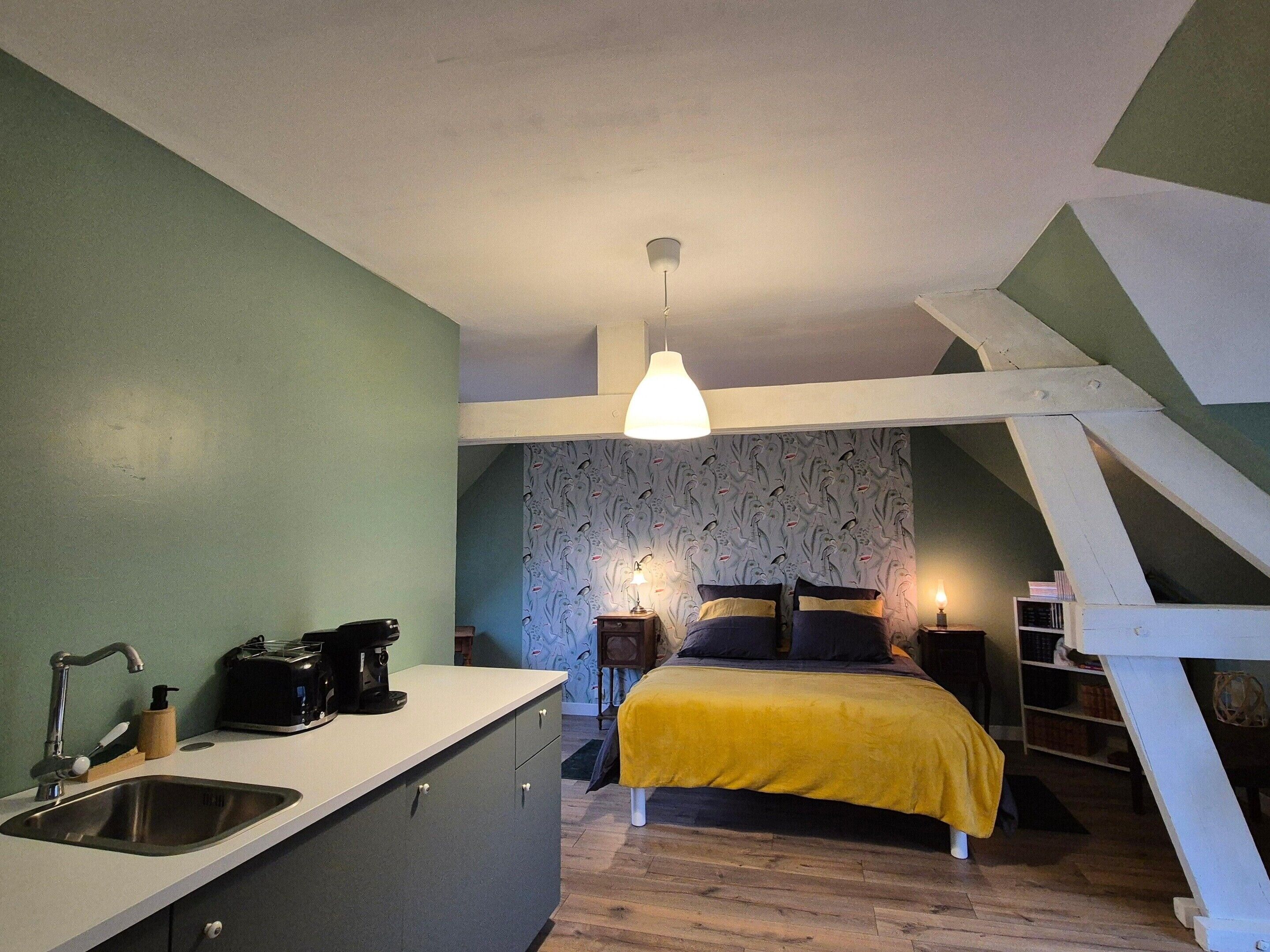 Quarto compacto com cama, mesa de cabeceira e um apoio para objetos. É visível uma área de cozinha com pia e torradeira.