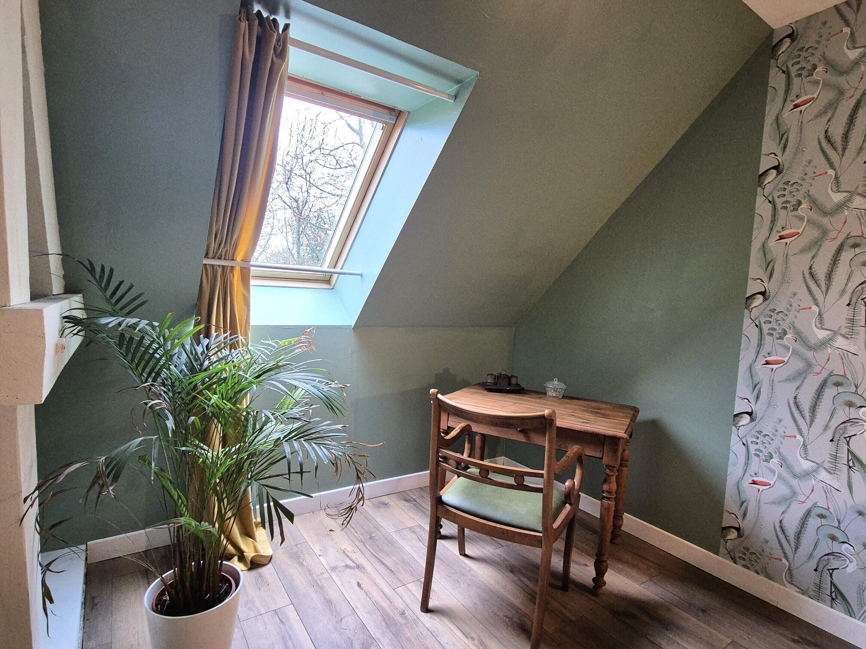Ein Zimmer mit schrägen Wänden, einem Holztisch mit Stuhl, einer Topfpflanze und einem Fenster mit Vorhängen.