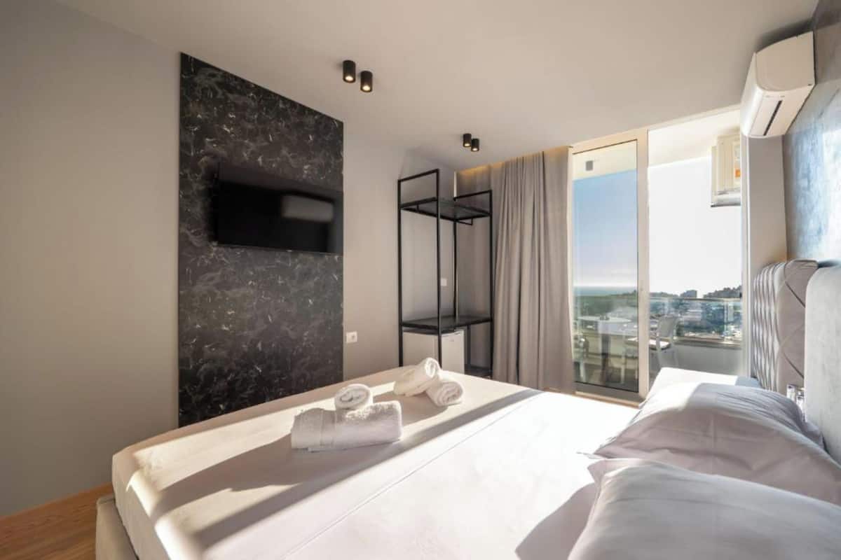 Una habitación de hotel moderna con una cama grande, un televisor de pantalla plana en la pared, un balcón con vistas a la ciudad y aire acondicionado integrado.