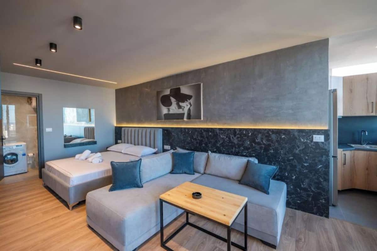 Una habitación de hotel moderna con sofá gris, una mesita de centro de madera y una pared decorativa de mármol.