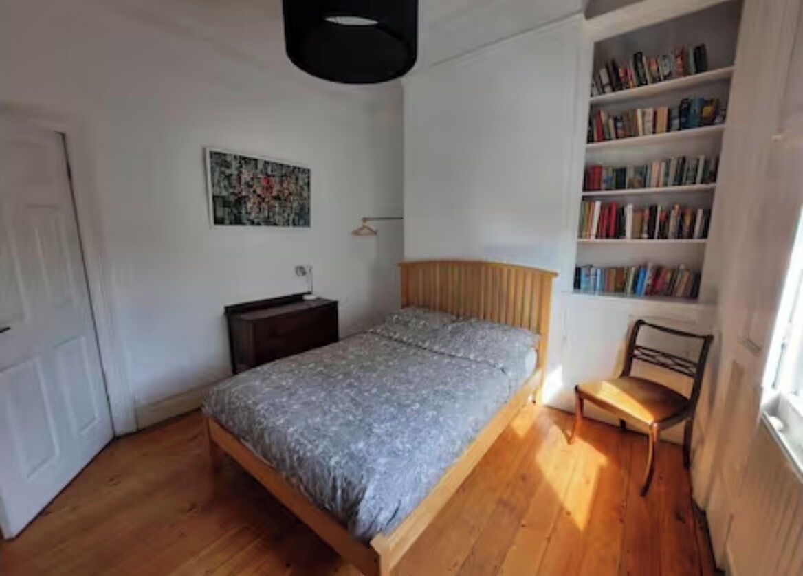 A bedroom with a wooden bed, a bedside table, a chair, a bookshelf, and a painting on the wall.