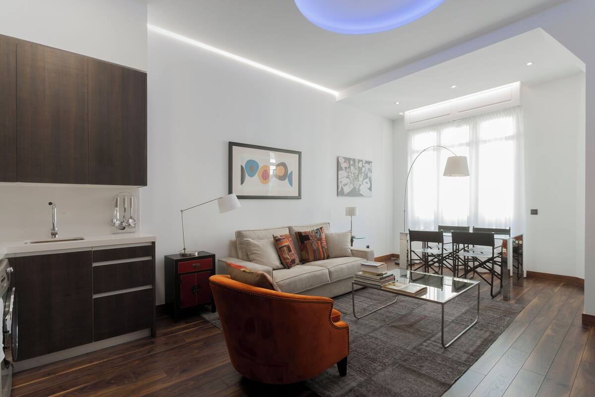 Een moderne woonkamer met een bank, salontafel en een oranje fauteuil. Er is een eethoek met stoelen en een tafel. De kamer heeft een groot raam met gordijnen en een plafondlamp.