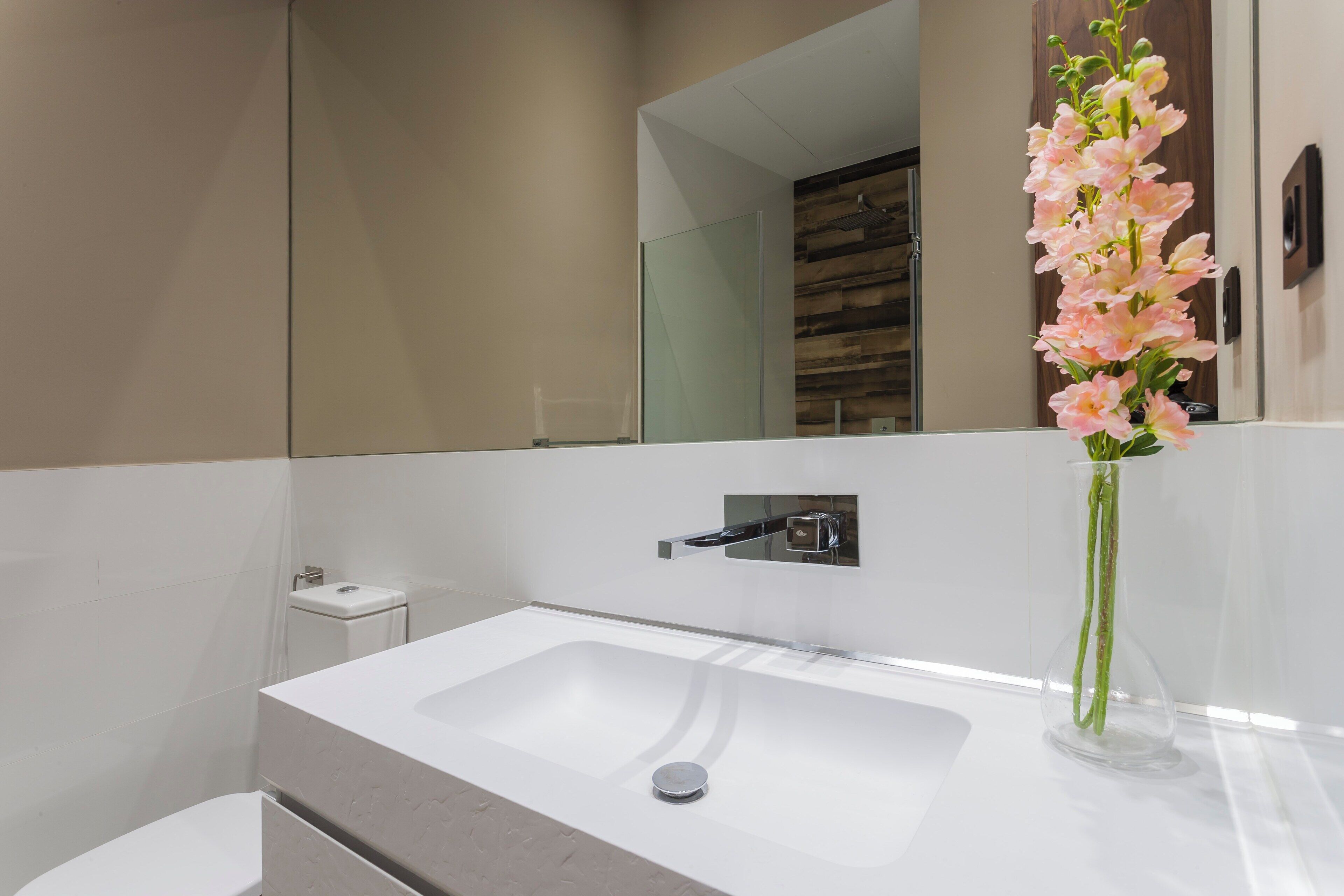 Un baño moderno con un lavabo blanco, un espejo y un florero con flores rosadas.