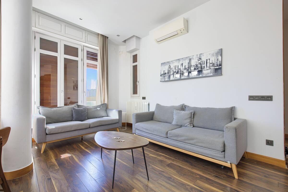 Een moderne woonkamer met twee grijze banken, een klein rond tafeltje en een stadsgezicht schilderij aan de muur.
