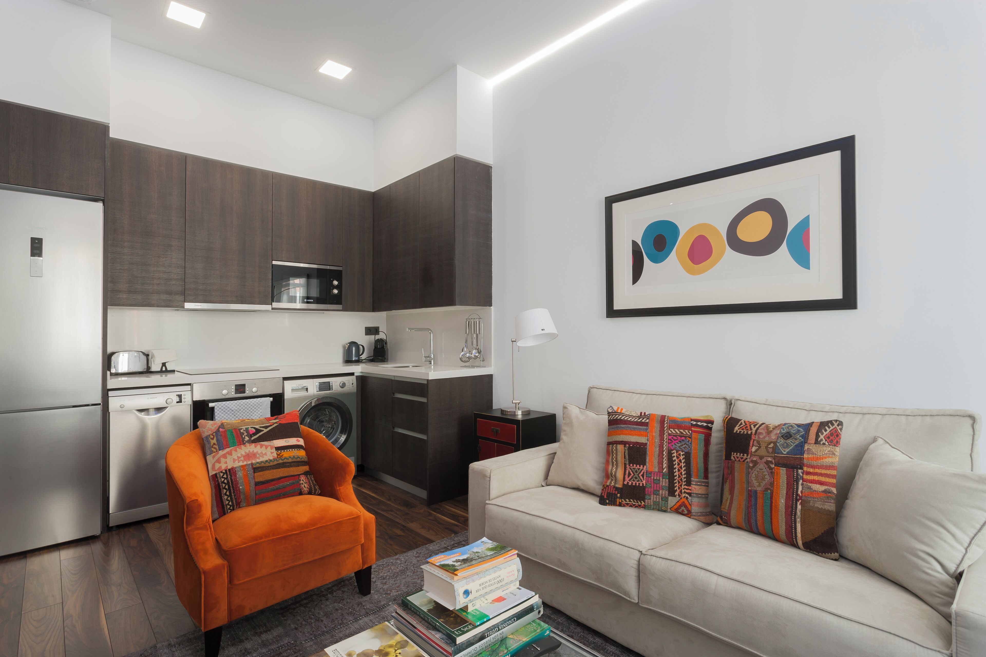 Una cocina moderna con armarios de madera oscura, un microondas y una cafetera. Hay un sofá con almohadones coloridos y un cuadro abstracto enmarcado en la pared.