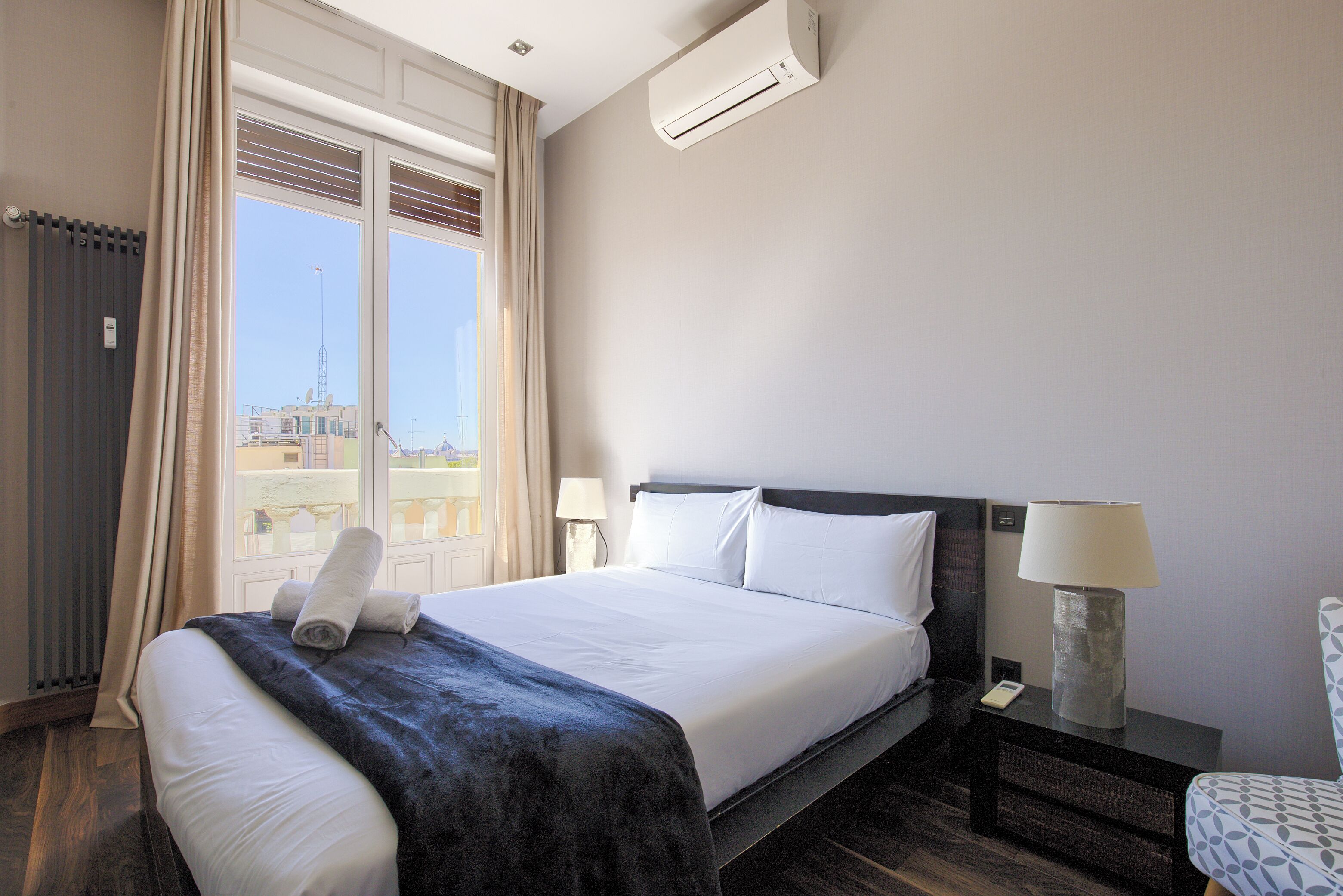 Habitación de hotel con una cama grande, una mesita de noche con lámpara y vistas a edificios desde la ventana.