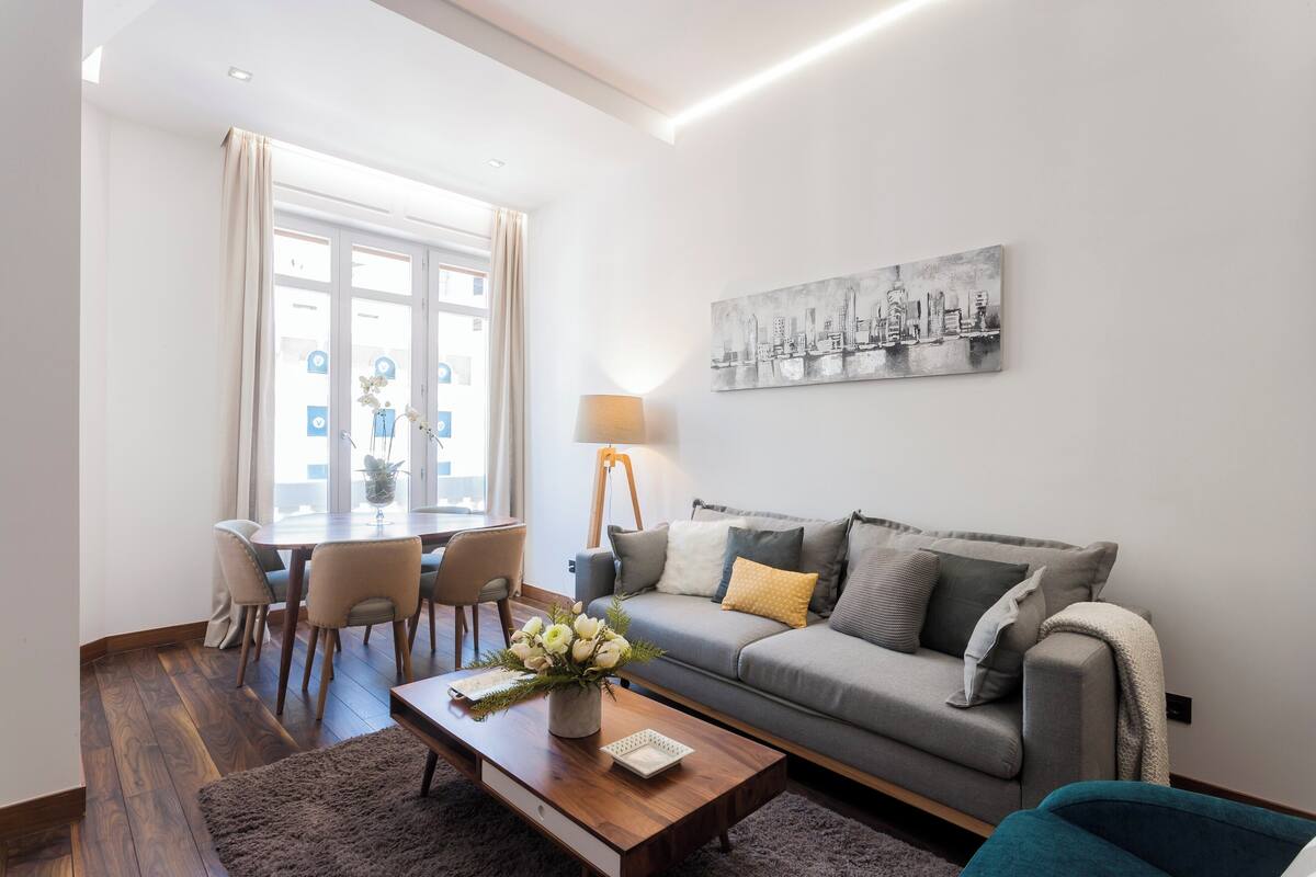 Een moderne woonkamer met een grijze bank, een houten salontafel en een eethoek met een tafel en stoelen.