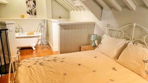 A bedroom with a bed, a nightstand, a lamp, a window, and a bathroom with a bathtub and a towel rack. - Cotswolds Country Cottage | Boutique, Romantic Retreat for Two (Purton Stoke)